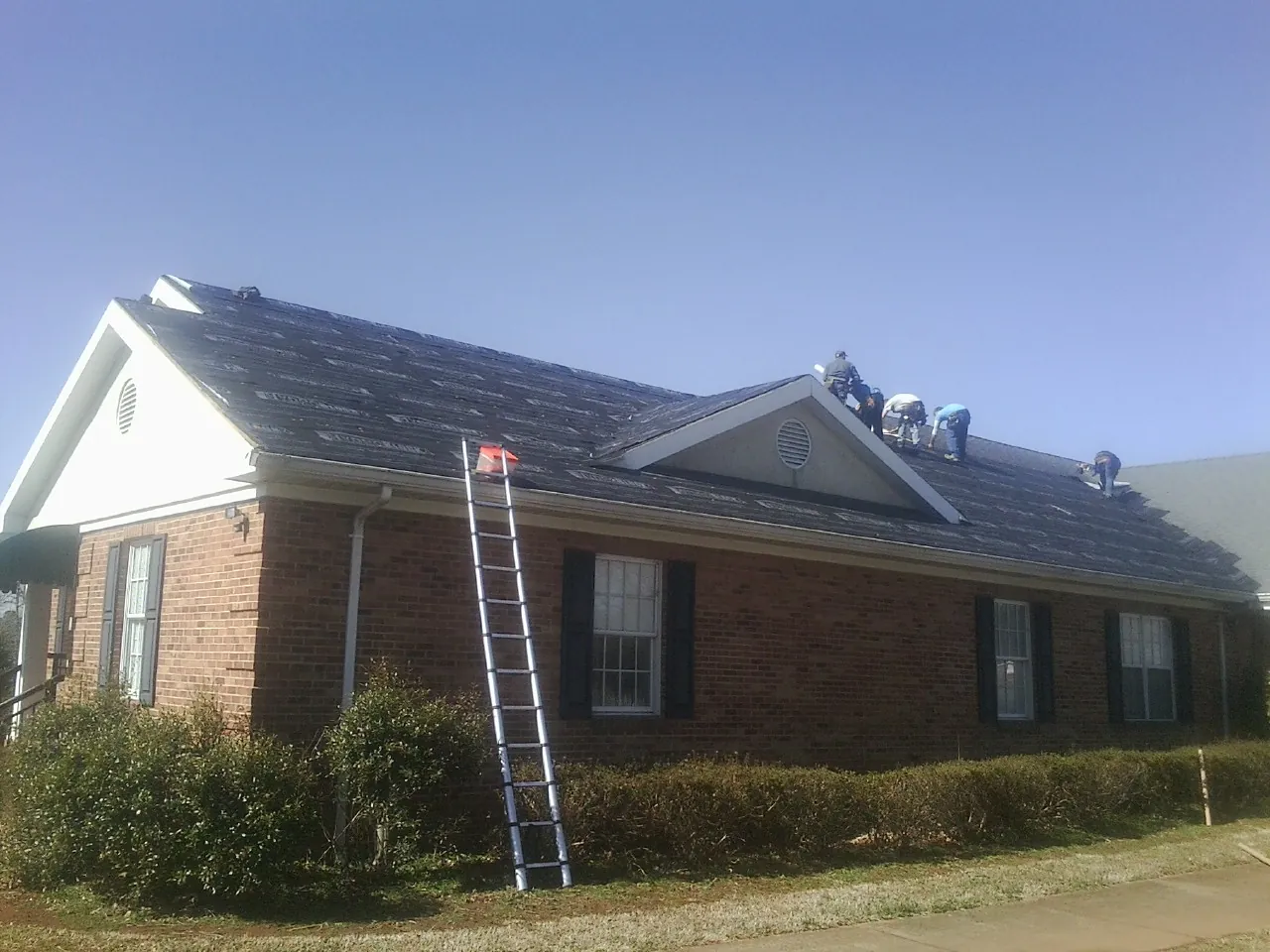 Crew tearing shingles off a brick ranch roof with a ladder set — Emergency Roof Repair work by our crew in Lenox